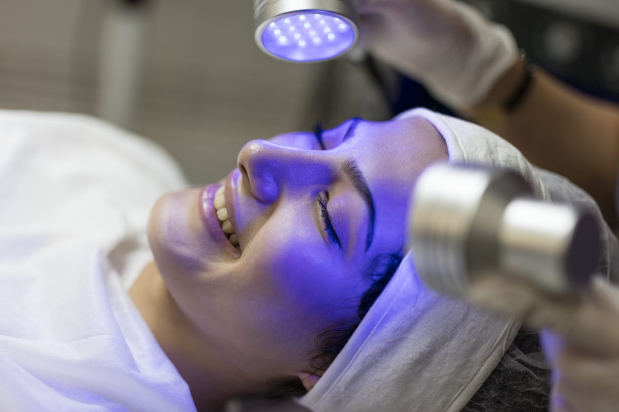 Female Beautician Doing Blue Light Therapy On The Face Of A Young Woman